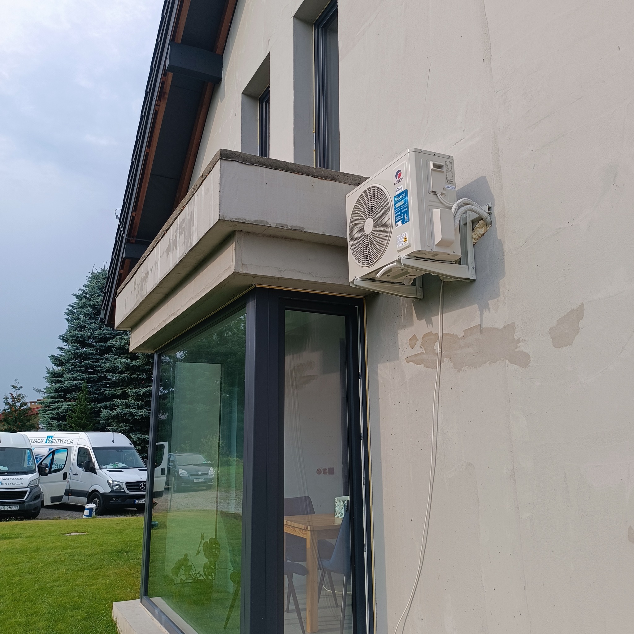 Zewnętrzna jednostka klimatyzacji marki Gree zamontowana na elewacji domu z widocznymi przewodami i pianką montażową, na tle zaparkowane busy z logo firmy instalacyjnej.