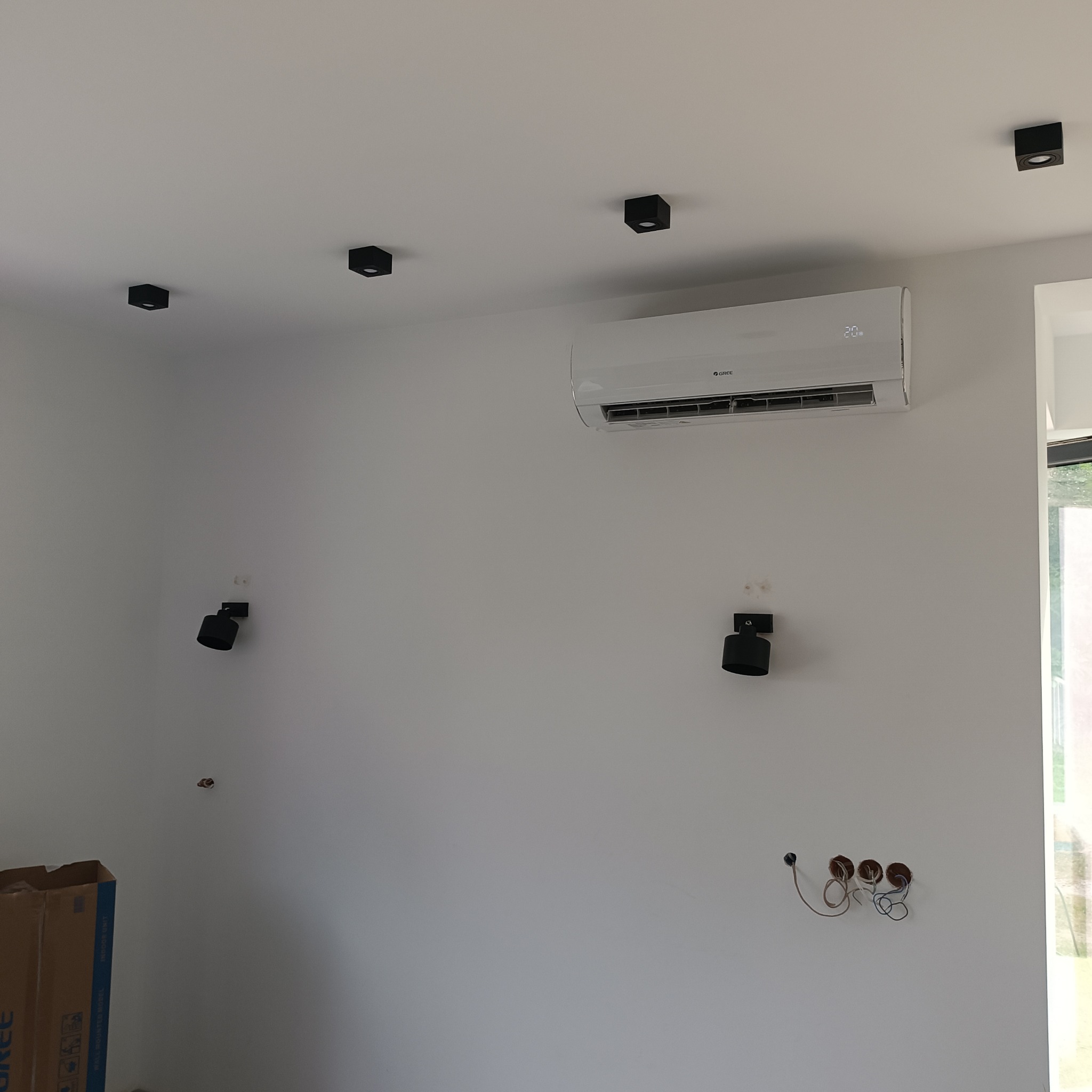 Wnętrze pokoju z zamontowanym klimatyzatorem marki Gree, widoczne czarne, kwadratowe lampy sufitowe i kinkiety na ścianie, instalacja elektryczna w ścianie, kartonowe pudło w rogu pokoju.
