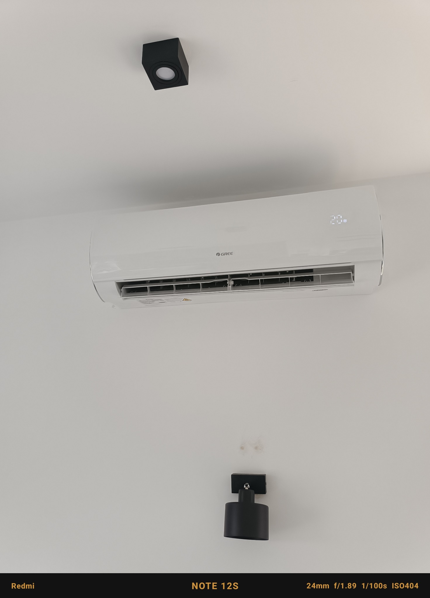 Biała klimatyzacja Gree zamontowana na białej ścianie, z czarnymi lampami sufitowymi i plamami na ścianie.