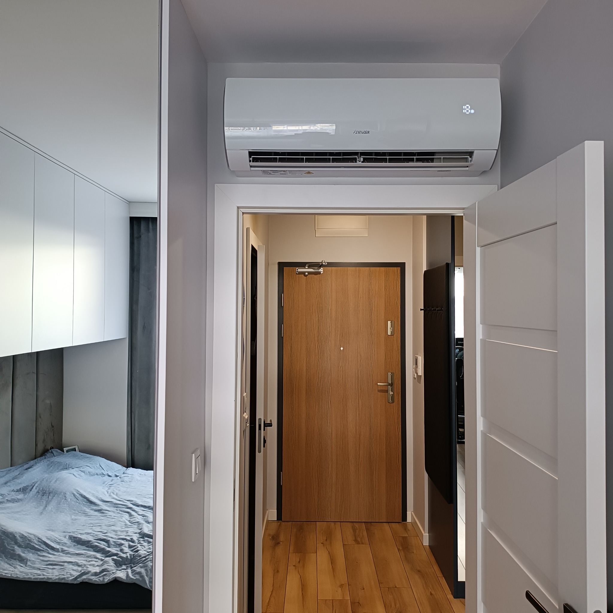 Biała jednostka klimatyzacyjna marki Sinclair zamontowana nad drzwiami w nowoczesnym mieszkaniu z widokiem na sypialnię i przedpokój z drewnianą podłogą.