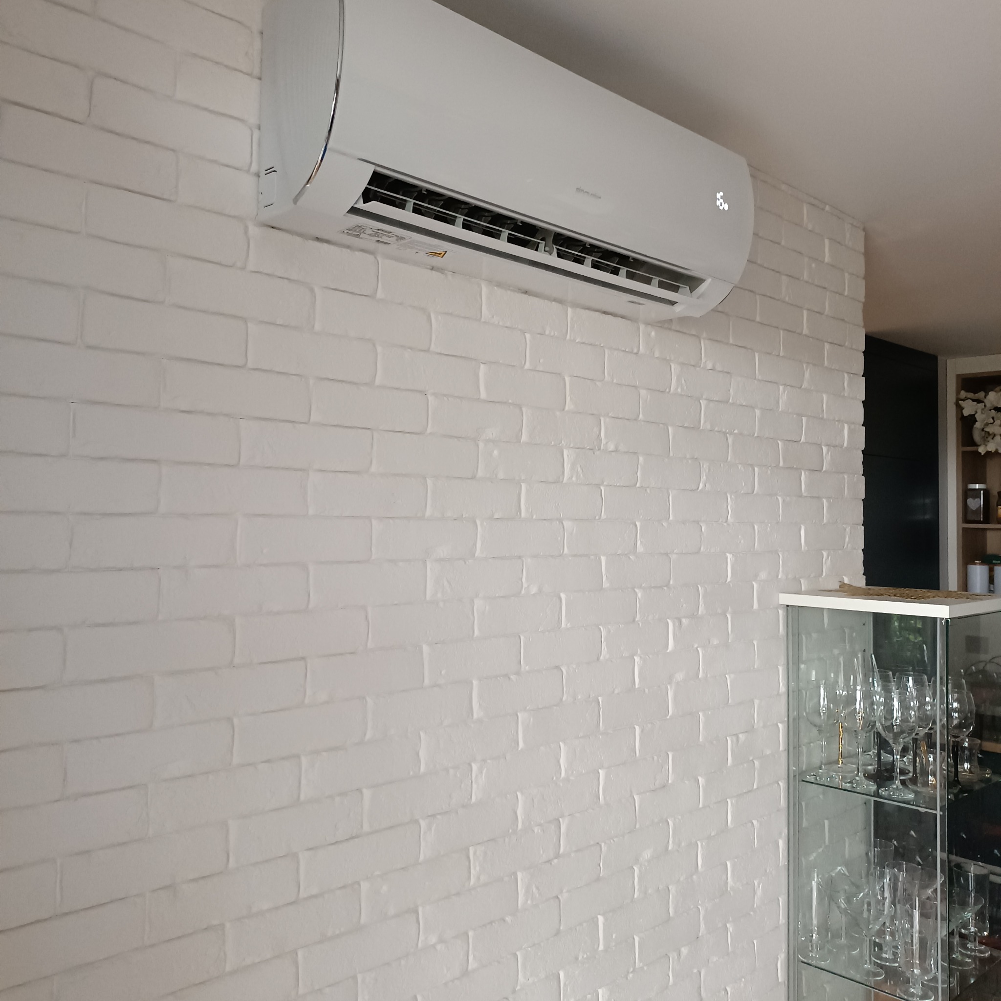Biały klimatyzator ścienny zamontowany na ścianie z białej cegły, obok szklana witryna z kieliszkami do wina.