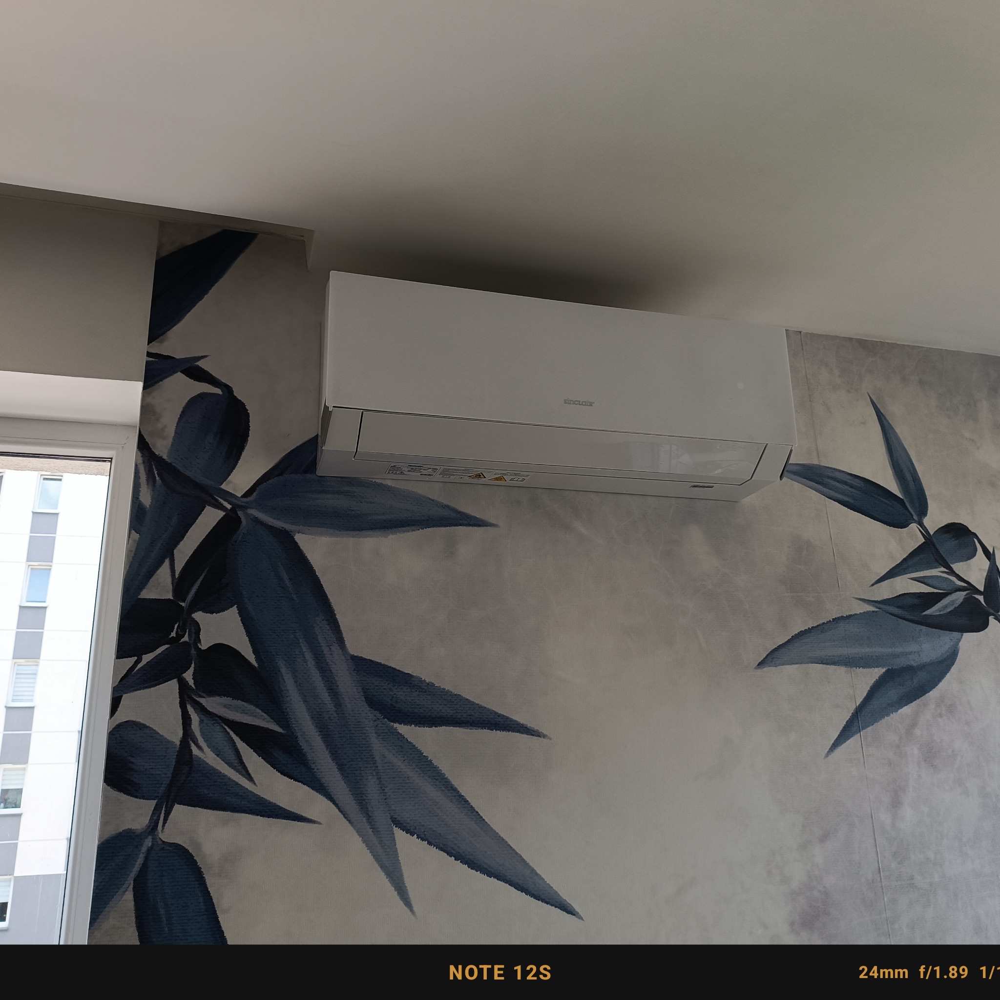 Biała jednostka klimatyzacji Sinclair zamontowana na ścianie z wzorem bambusowych liści w odcieniach szarości i granatu, widoczna część okna po lewej stronie.