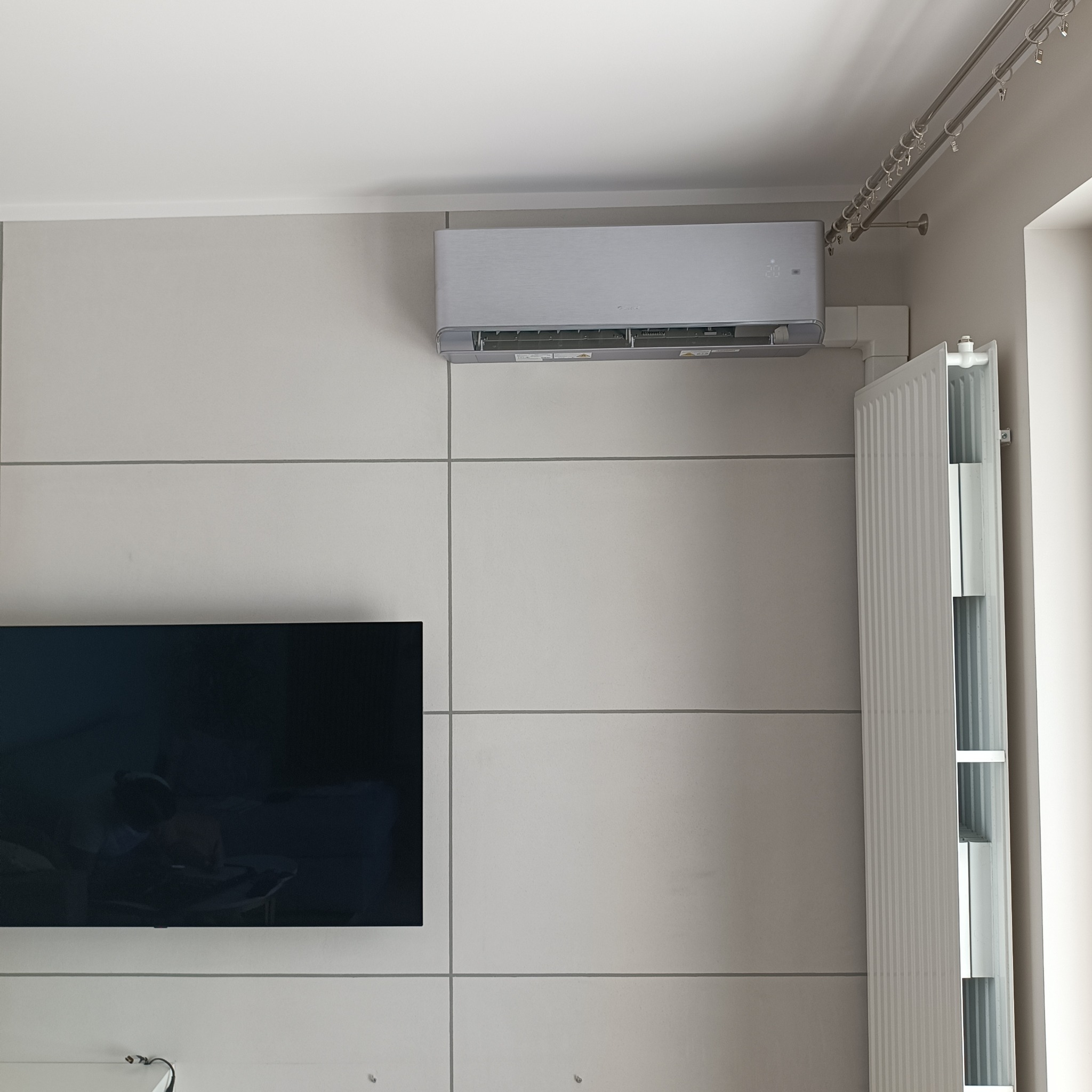 Zamontowany srebrny klimatyzator ścienny w nowoczesnym wnętrzu z panelem ściennym, obok grzejnika i telewizora.