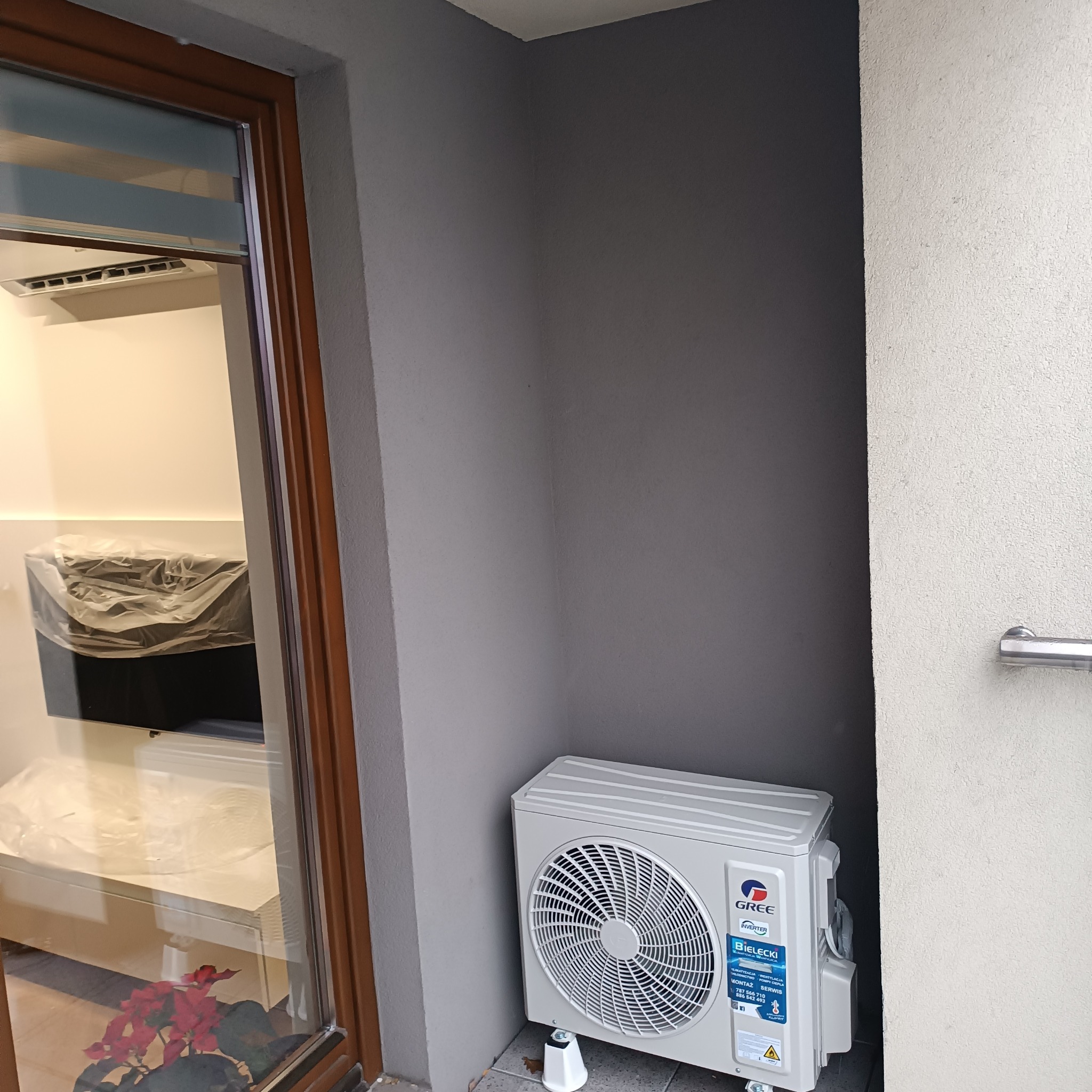 Zewnętrzna jednostka klimatyzacji Gree zamontowana przy ścianie budynku, widoczna z boku okna z kwiatami w doniczce.