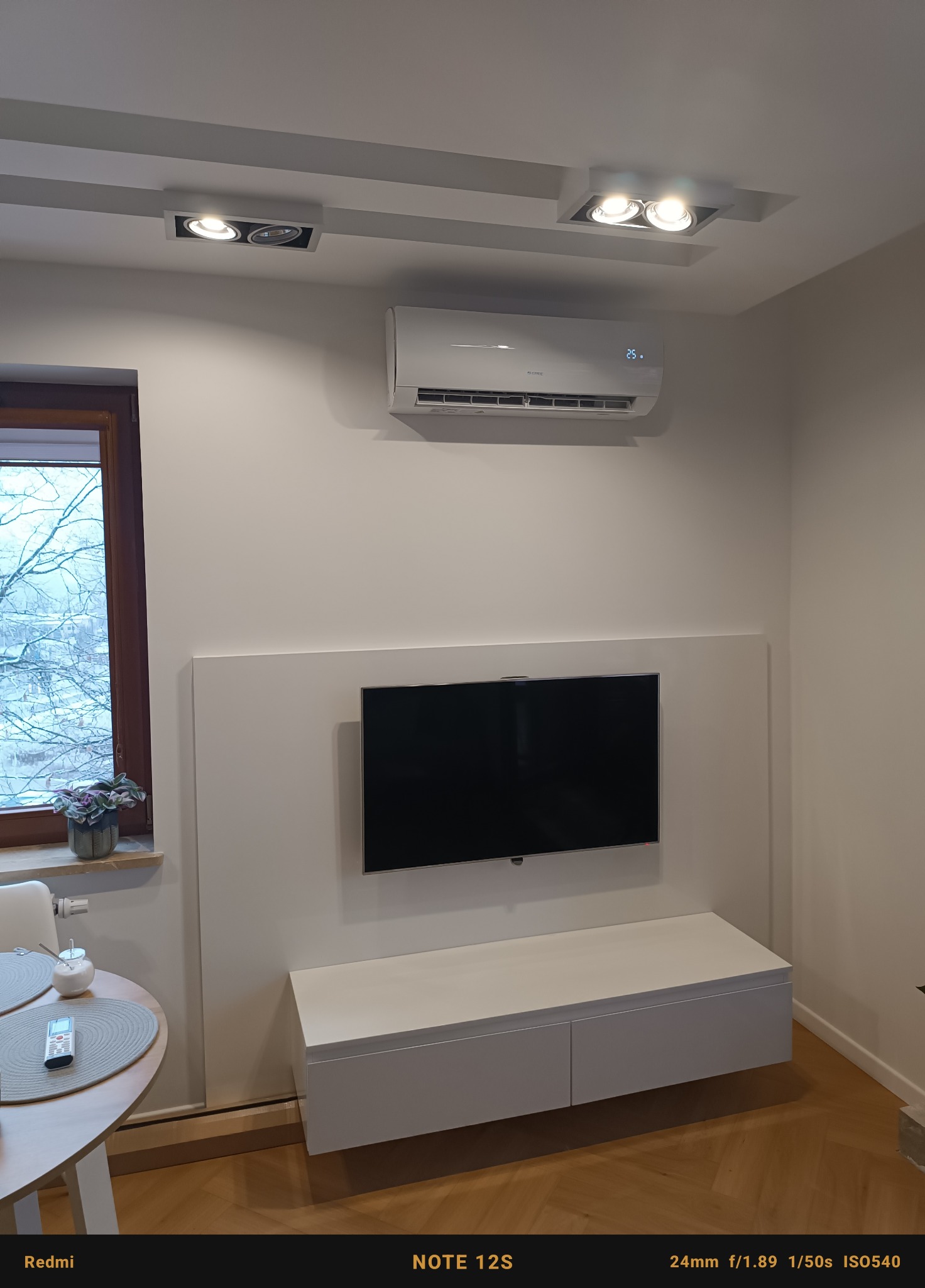 Biały klimatyzator ścienny z wyświetlaczem temperatury 25 stopni Celsjusza, zamontowany nad telewizorem w nowoczesnym salonie z parkietem i oknem.