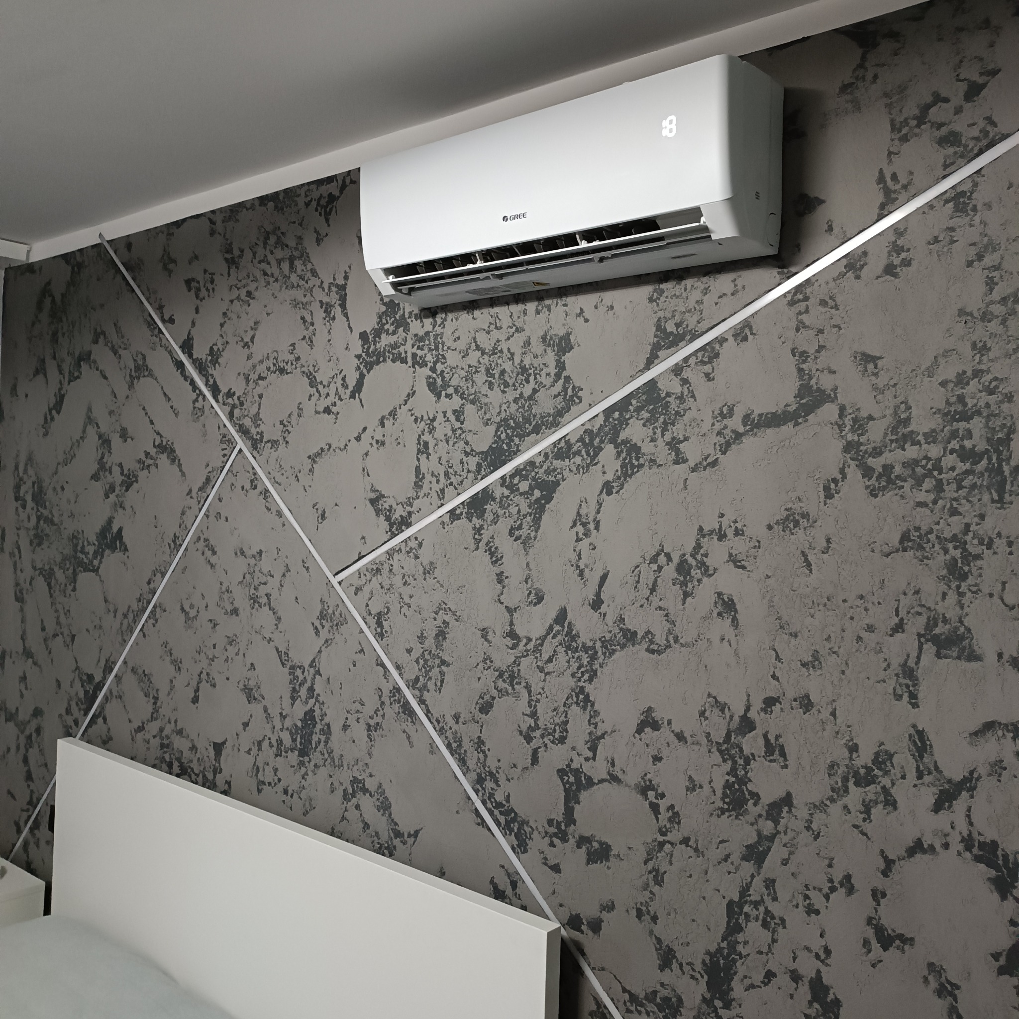Biały klimatyzator marki Gree zamontowany na szarej, teksturowanej ścianie z dekoracyjnymi białymi listwami, widoczny fragment białego zagłówka łóżka.