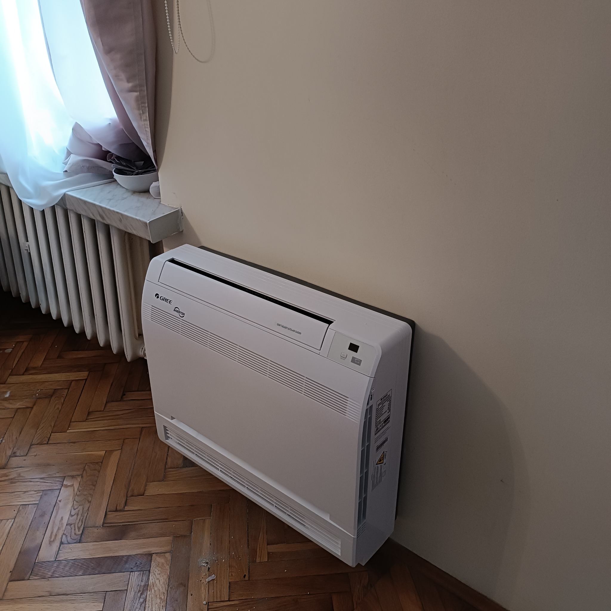 Biała, podłogowa jednostka klimatyzacji marki Gree, umieszczona w rogu pokoju przy ścianie, obok grzejnika i okna z zasłonami, na parkiecie ułożonym w jodełkę.