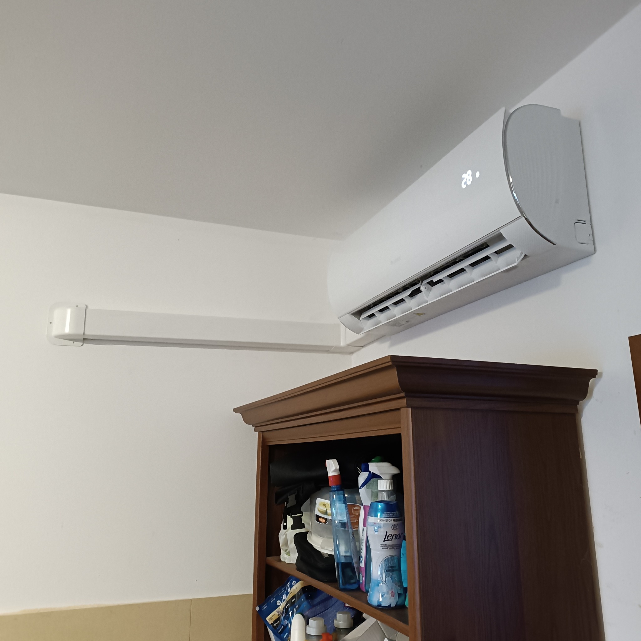 Biały klimatyzator ścienny z wyświetlaną temperaturą 28 stopni Celsjusza, zamontowany w pomieszczeniu nad ciemnobrązowym regałem z różnymi przedmiotami.