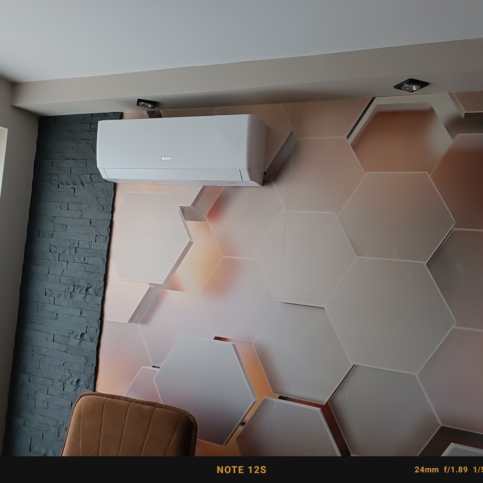 Biały klimatyzator marki Gree zamontowany na ścianie z dekoracyjną tapetą w kształcie plastra miodu, obok ściany z szarej cegły dekoracyjnej, widoczny fragment brązowego krzesła.