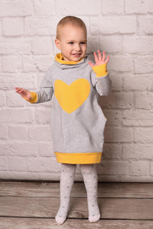 Uśmiechnięta dziewczynka w szarej sukience dresowej z żółtym sercem, żółtym golfem i mankietami, oraz białych rajstopach w serduszka, pozuje na tle ściany z białej cegły i drewnianej podłogi.