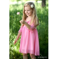 Uśmiechnięta dziewczynka w różowej, zwiewnej sukience z krótkim rękawem, trzymająca pojedynczy kwiat na tle zielonej trawy i drzew.