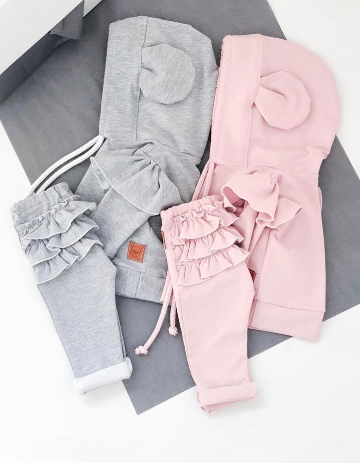 Dwa komplety dziecięce: szary i różowy, bluzy z kapturem ozdobionym uszkami i spodnie z falbankami, leżą na szaro-białym tle.