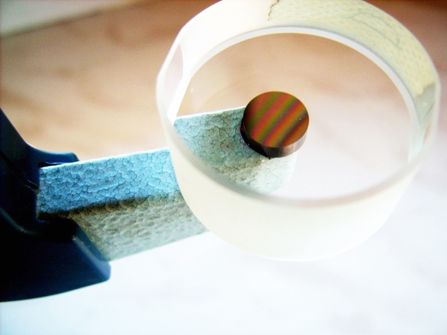 Miernik grubości powłok z okrągłą próbką z tęczowymi refleksami, umieszczoną w uchwycie.