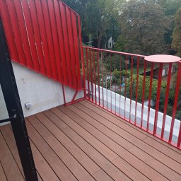 ANPOL - Czerwona balustrada z metalowych prętów na balkonie z drewnianą podłogą, widok na zieleń drzew.