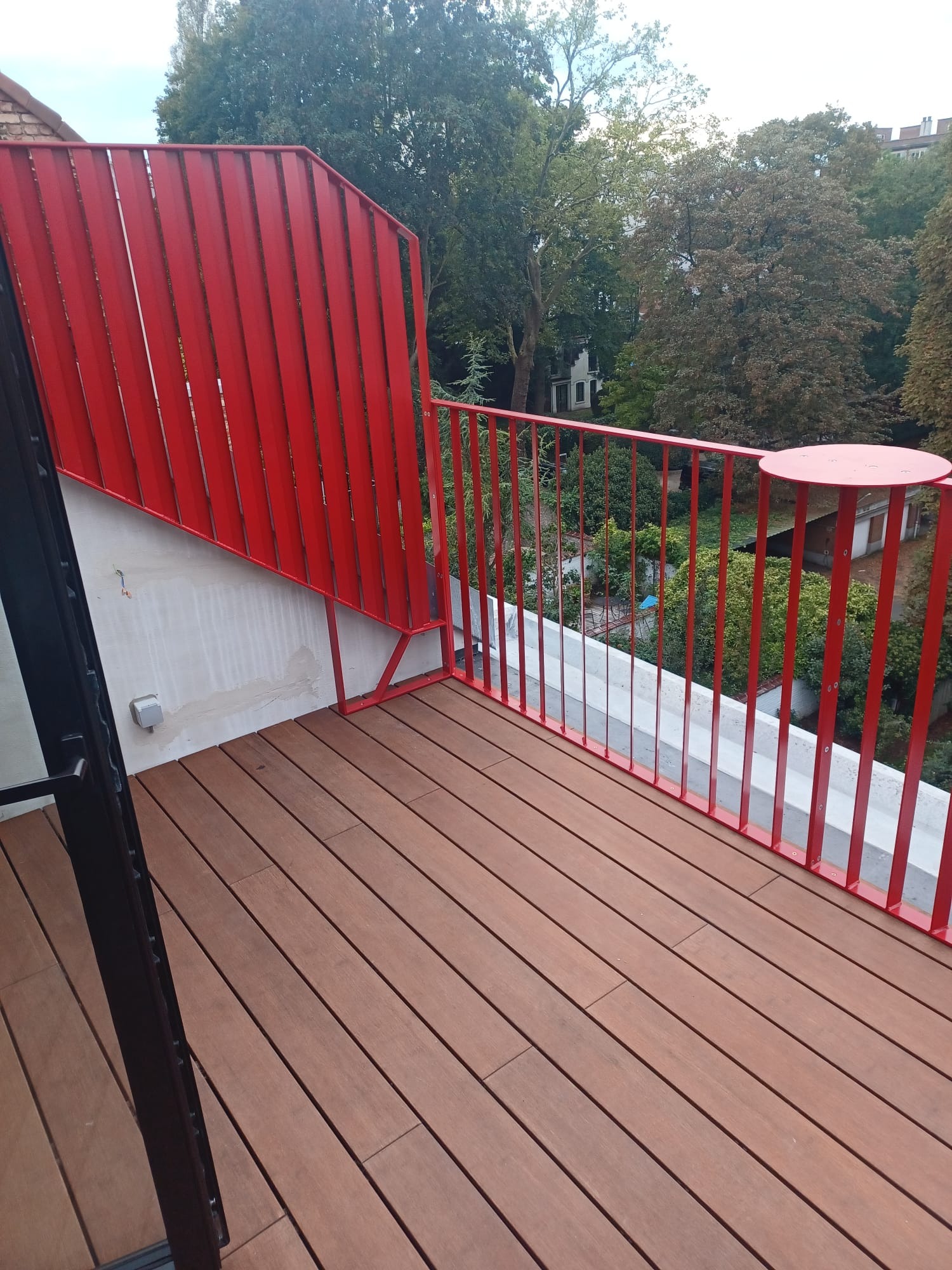 Czerwona balustrada z metalowych prętów na balkonie z drewnianą podłogą, widok na zieleń drzew.