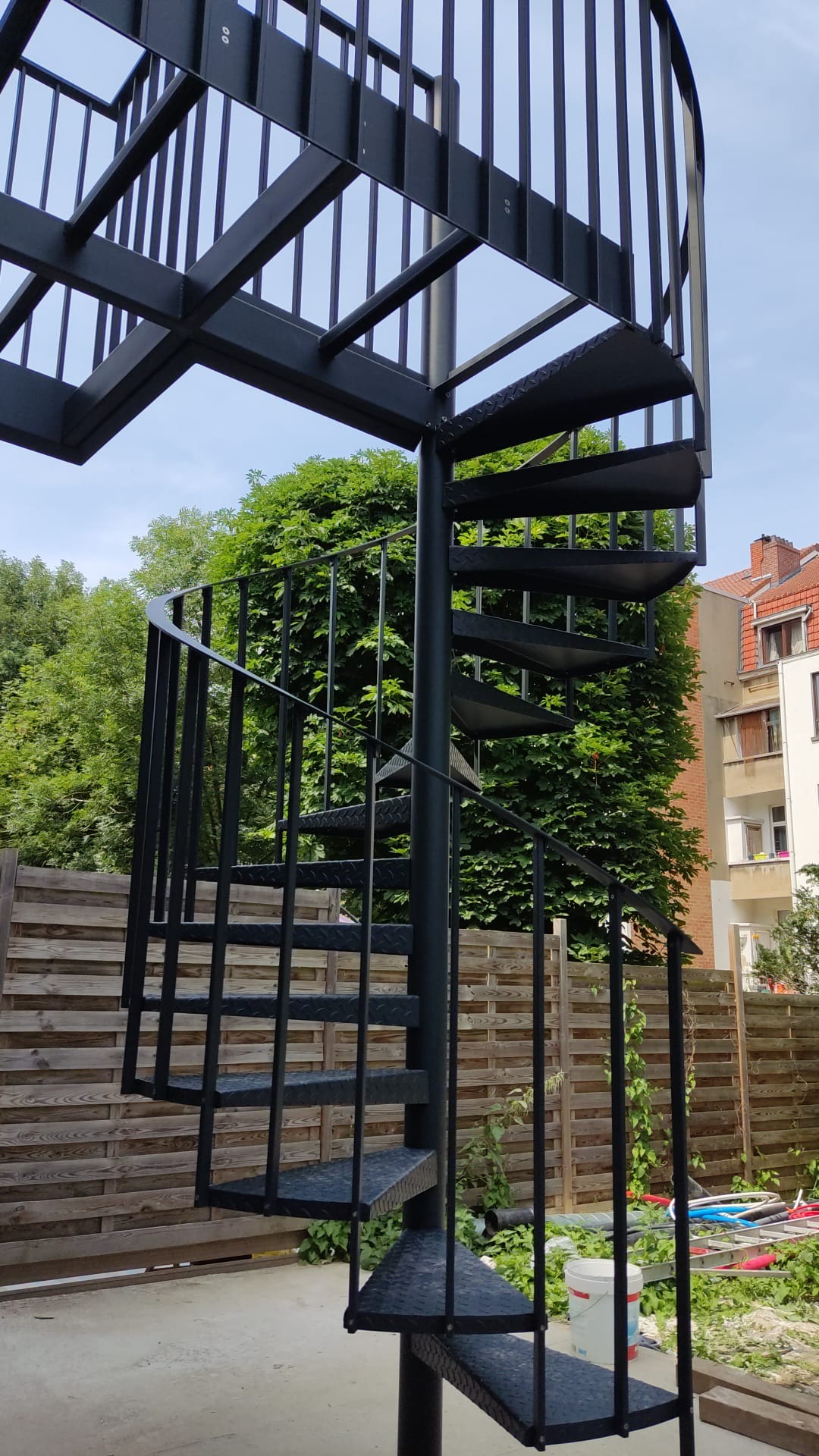 Metalowe schody spiralne zewnętrzne, pomalowane na czarno, z ażurową balustradą, na tle drewnianego ogrodzenia i zieleni.