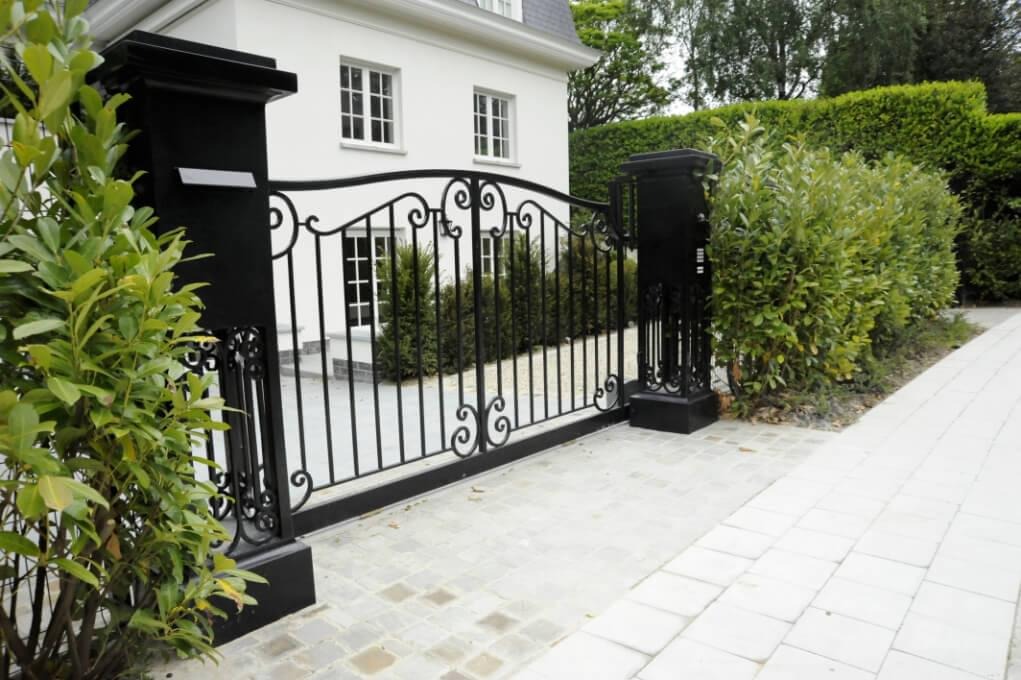 Elegancka, czarna brama wjazdowa z kutymi zdobieniami, zamontowana na posesji z białym domem i starannie przystrzyżonym żywopłotem.