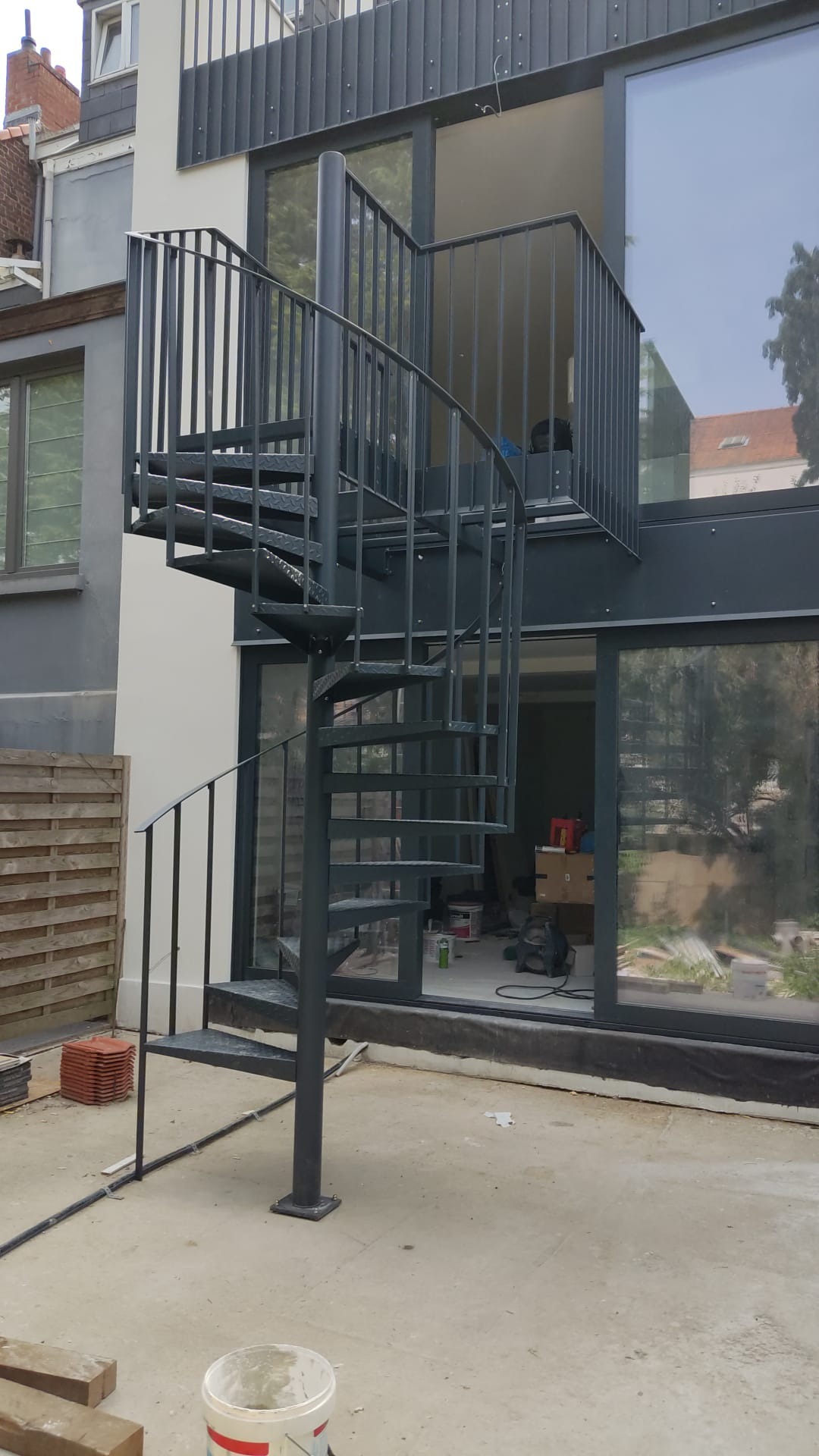 Zewnętrzne metalowe schody spiralne prowadzące na balkon, z widocznym fragmentem drewnianego ogrodzenia i elementami wykończeniowymi na ziemi.
