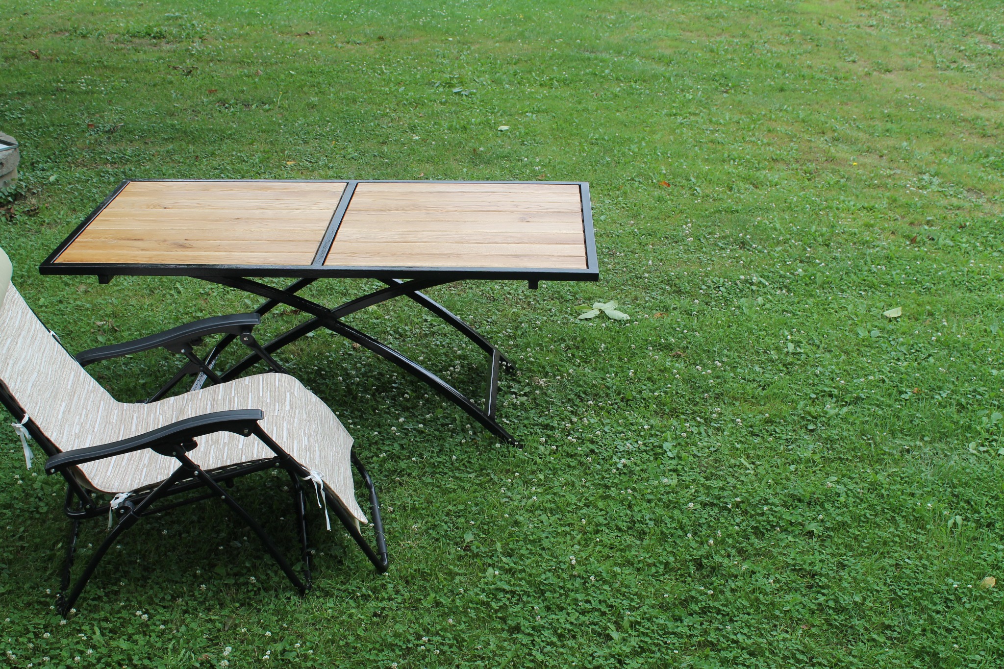 Składany stół ogrodowy z drewnianym blatem i metalową ramą oraz składane krzesło na trawniku z koniczyną.