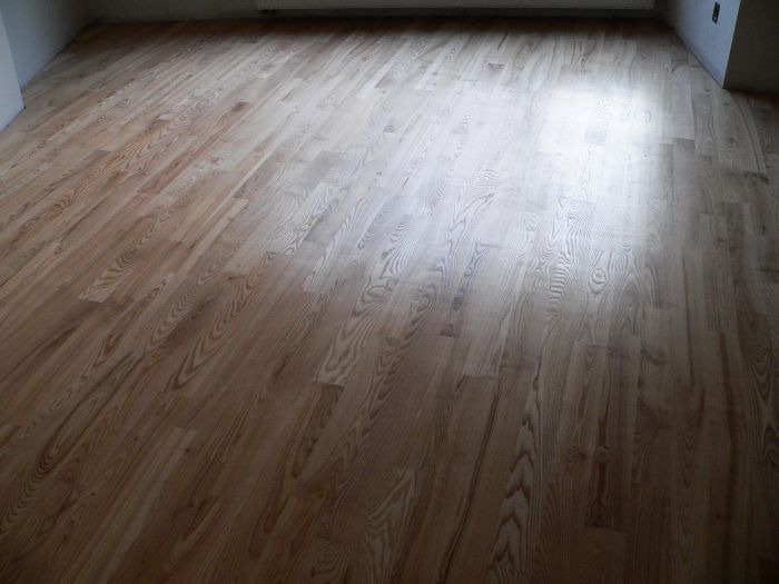 Ułożony parkiet drewniany z widocznym usłojeniem i delikatnym połyskiem, oświetlony naturalnym światłem wpadającym z okna, w tle ściana z grzejnikiem.