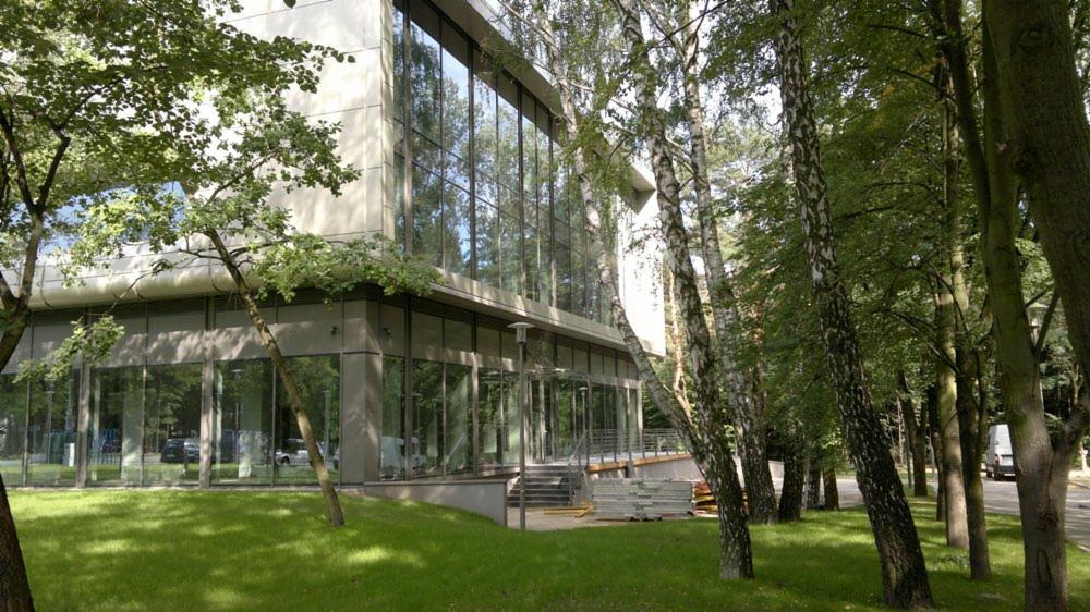 Nowoczesny budynek biurowy z dużą szklaną fasadą, otoczony zielenią i drzewami, widok z zewnątrz na trawnik i częściowo widoczne materiały budowlane przed wejściem.