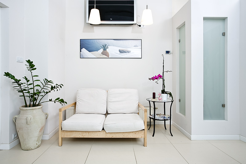 Jasne wnętrze z drewnianą sofą z białymi poduszkami, dużą donicą z zieloną rośliną, stolikiem z orchideą i obrazem nad sofą.