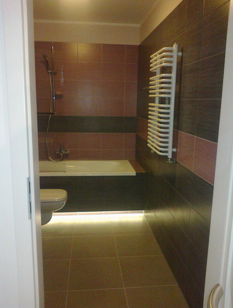 Wykończona łazienka z brązowymi płytkami, podświetleniem LED pod wanną i białym grzejnikiem drabinkowym. Widok zza uchylonych drzwi.