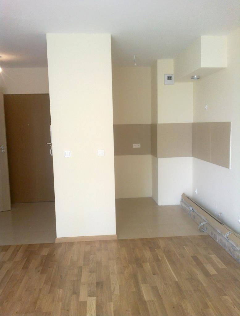 Jasne wnętrze po odświeżeniu: parkiet, kremowe ściany z beżowymi akcentami w kuchni, widoczne gniazdka i włączniki, listwy przypodłogowe i drzwi wejściowe.