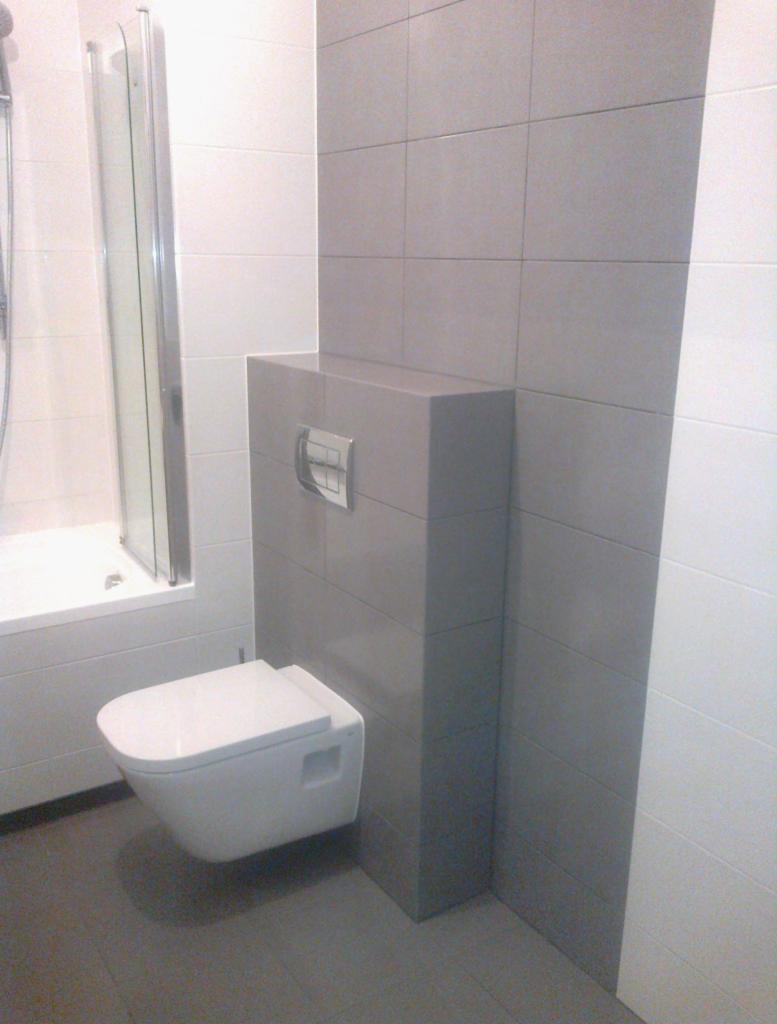 Nowoczesna toaleta podwieszana w szaro-białej łazience z kabiną prysznicową. Minimalistyczny design, płytki w odcieniach szarości, chromowany przycisk spłukiwania.