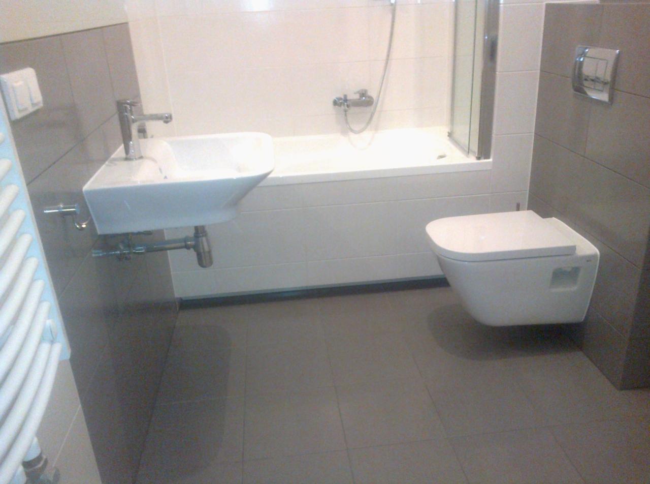 Nowoczesna łazienka po remoncie: biała armatura (umywalka, toaleta) na tle szarych płytek. Widoczna wanna z parawanem i grzejnik.