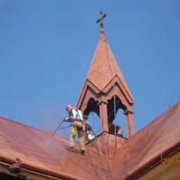 Pracownik w uprzęży zabezpieczającej czyści dach kościoła z wieżyczką i krzyżem za pomocą myjki ciśnieniowej, widoczny efekt czyszczenia, data na zdjęciu wskazuje na rok 2010.
