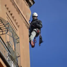 Pracownik na linie zabezpieczonej do ściany budynku, w kasku i uprzęży, wykonujący prace wysokościowe przy elewacji z oknem z kratą.