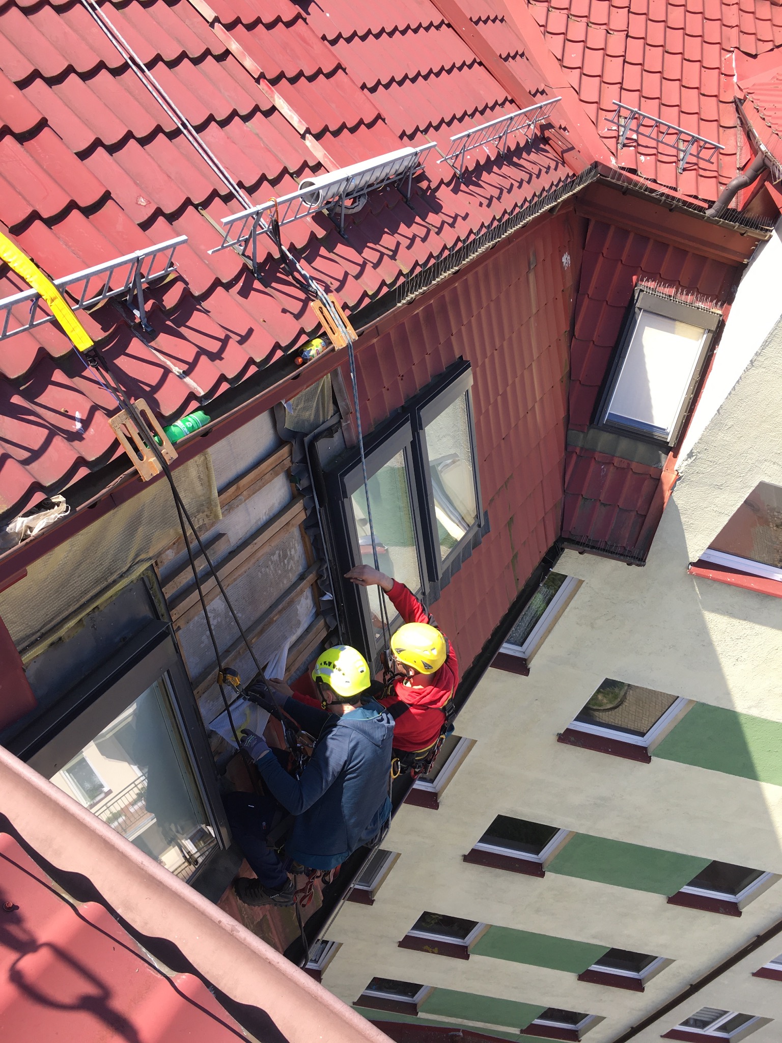 Dwóch pracowników w kaskach, zabezpieczonych linami, wykonuje prace na wysokości przy ścianie budynku z czerwonym dachem, widoczne rusztowania i okna dachowe.