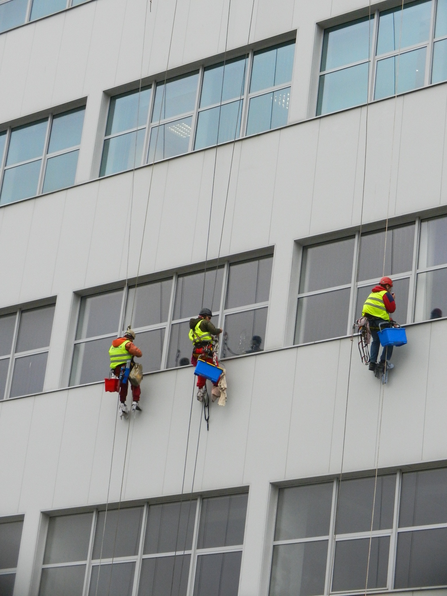Trzech alpinistów przemysłowych na linach czyści okna biurowca, każdy z nich ma inny kolor kasku i akcesoriów, a na elewacji widoczne są smugi po czyszczeniu.