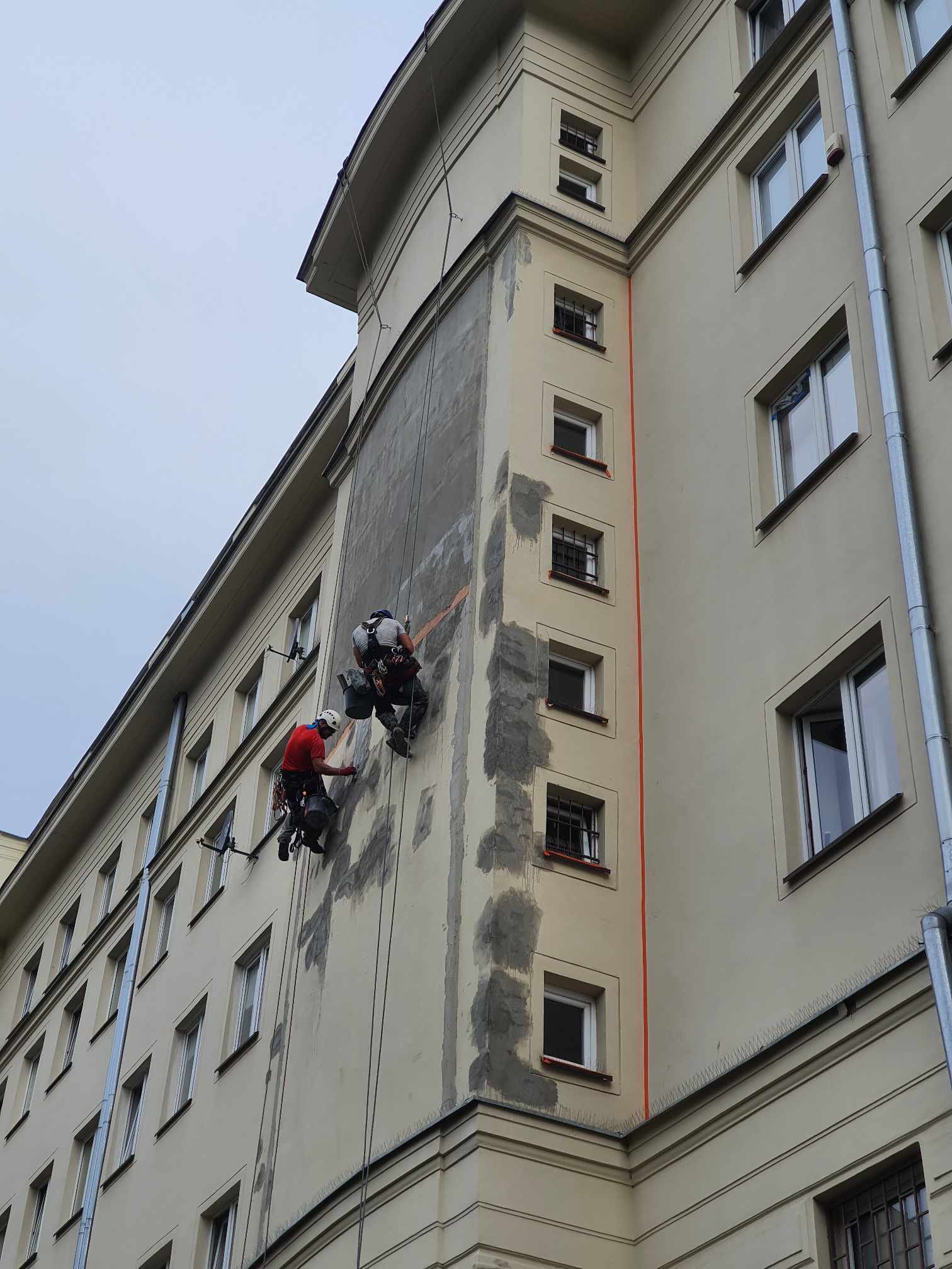 Dwóch pracowników na linach zabezpieczających wykonuje prace na elewacji wysokiego budynku, widoczne ślady po naprawach i przygotowaniu do malowania.