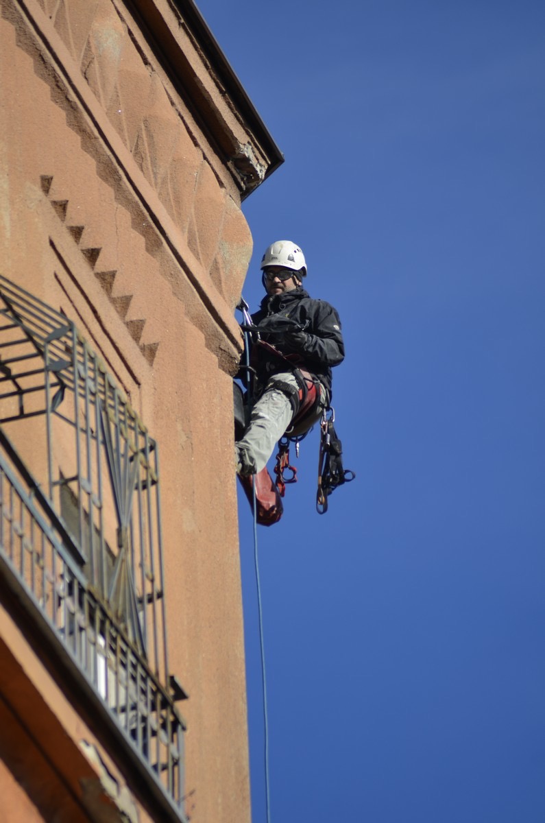 Pracownik na linie zabezpieczonej do ściany budynku, w kasku i uprzęży, wykonujący prace wysokościowe przy elewacji z oknem z kratą.