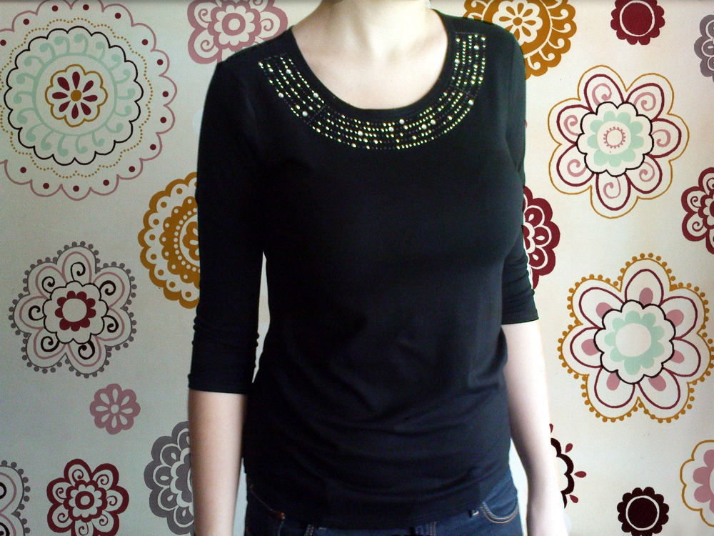 Czarna bluzka z rękawem 3/4, zdobiona aplikacją z dżetów i koralików przy dekolcie, na tle ściany w kwiatowe wzory.