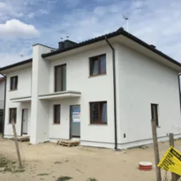 Biały, dwukondygnacyjny dom bliźniak z brązowymi oknami, z tablicą informacyjną budowy i paletami przy wejściu, na tle błękitnego nieba.