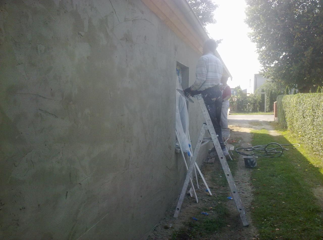 Pracownik na drabinie wyrównuje świeżo nałożony tynk na elewacji budynku jednorodzinnego, widoczne zacieki i nierówności na powierzchni ściany, w tle zieleń i ulica.