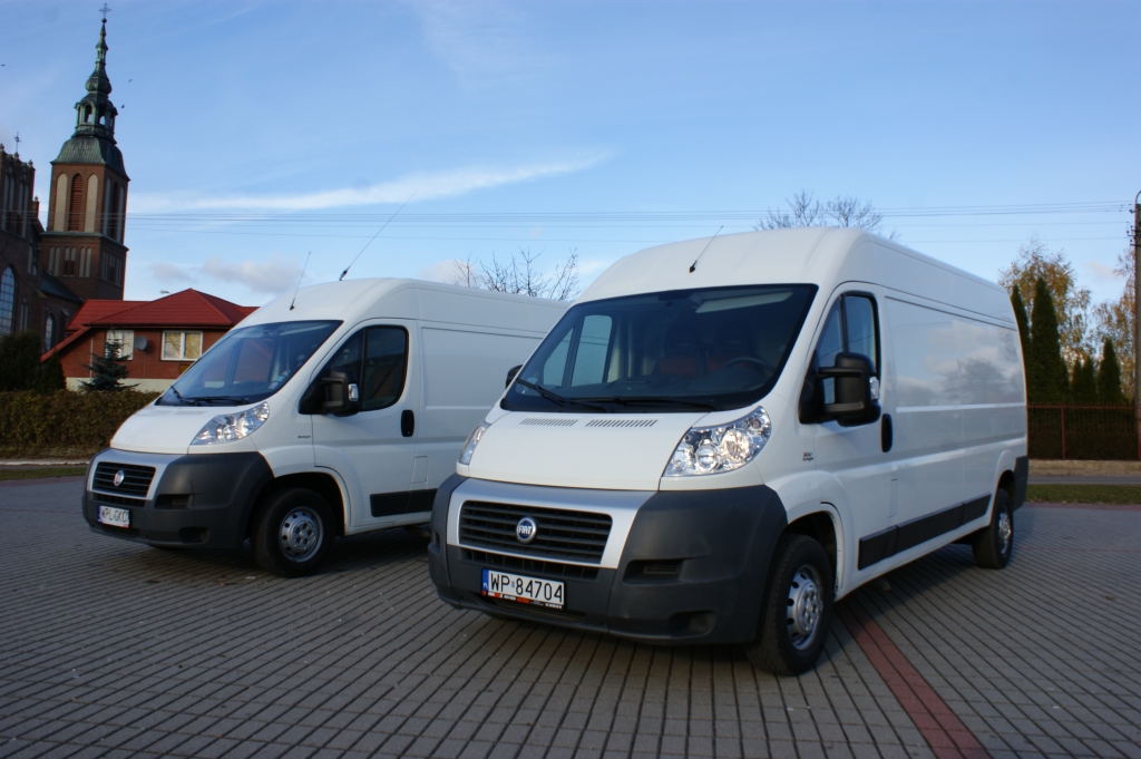 Dwa białe busy Fiat Ducato zaparkowane na kostce brukowej, w tle kościół z wysoką wieżą, błękitne niebo.