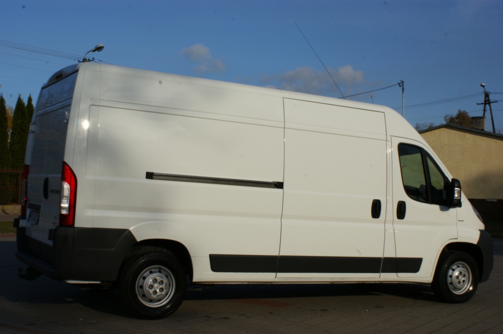 Biały samochód dostawczy z anteną na dachu, zaparkowany na kostce brukowej w słoneczny dzień, widok z boku.