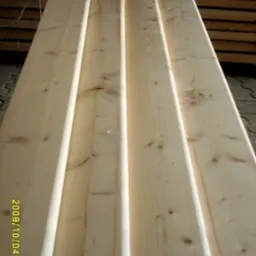 Stos jasnych, sosnowych desek elewacyjnych typu klin, ułożonych jedna na drugiej, widoczne sęki i naturalny rysunek drewna.