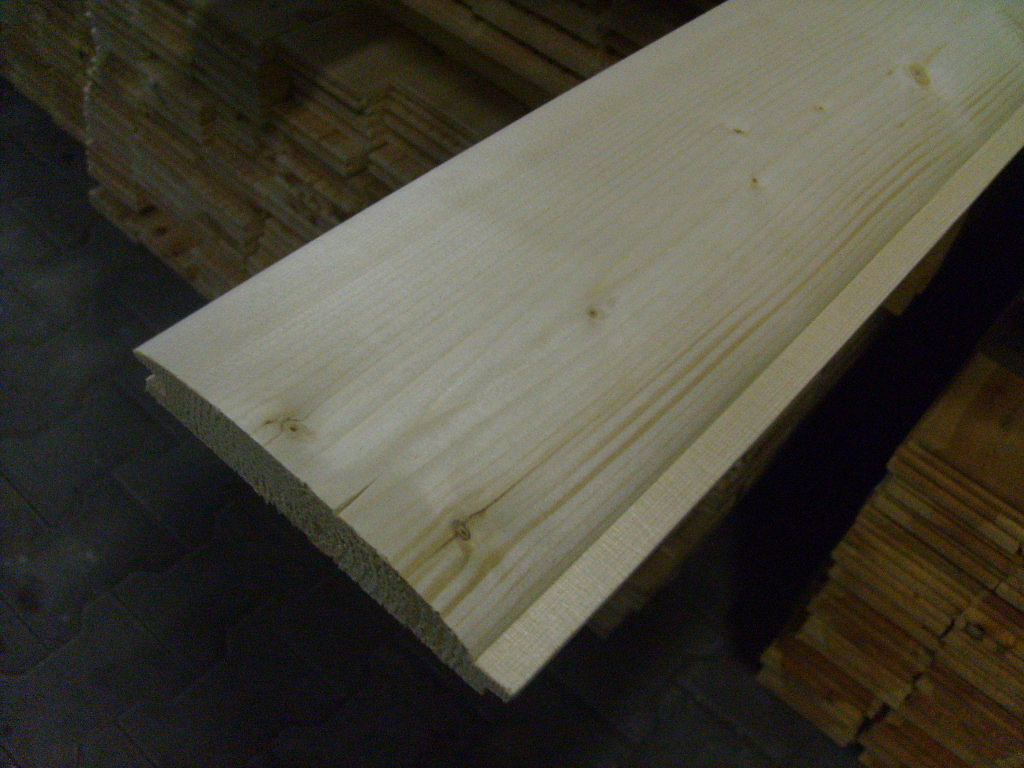 Profilowana deska z jasnego drewna z widocznymi słojami i sękami, ułożona na stosie innych desek w magazynie.