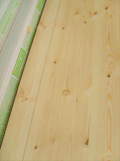 Jasne deski podłogowe z widocznymi sękami, ułożone pionowo, z fragmentem opakowania z logo producenta widocznym po lewej stronie.