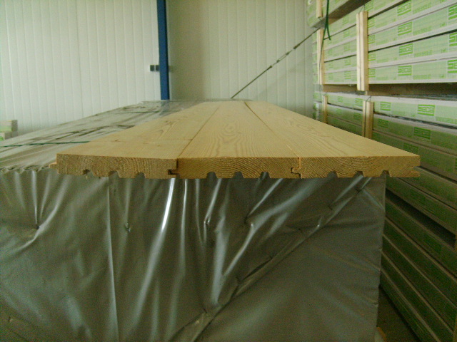 Profilowane deski podłogowe z jasnego drewna ułożone na palecie owiniętej folią, widoczne połączenie pióro-wpust, w tle magazyn z zapasem desek.
