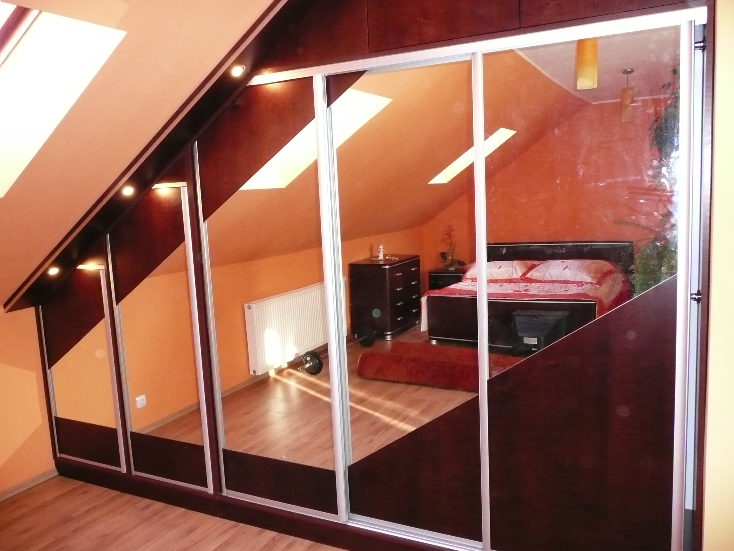 Szafa wnękowa z przesuwnymi lustrzanymi drzwiami na poddaszu, widoczna sypialnia z łóżkiem i komodą, drewniana podłoga, skosy sufitowe z oświetleniem punktowym.