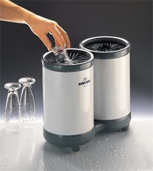 Mycie szklanki w profesjonalnej myjce do szkła; widoczne dwie myjki koloru białego z szarymi elementami na blacie z kroplami wody; w jednej myjce umieszczana jest szklanka przez dłoń; w tle dwie...