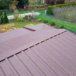 Widok z góry na brązowy dach z blachy trapezowej, pokryty kroplami wody po deszczu, z widocznym ogrodem w tle.