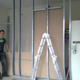 Montaż płyty karton gips na metalowym stelażu, widoczny pracownik w kombinezonie i drabina aluminiowa.