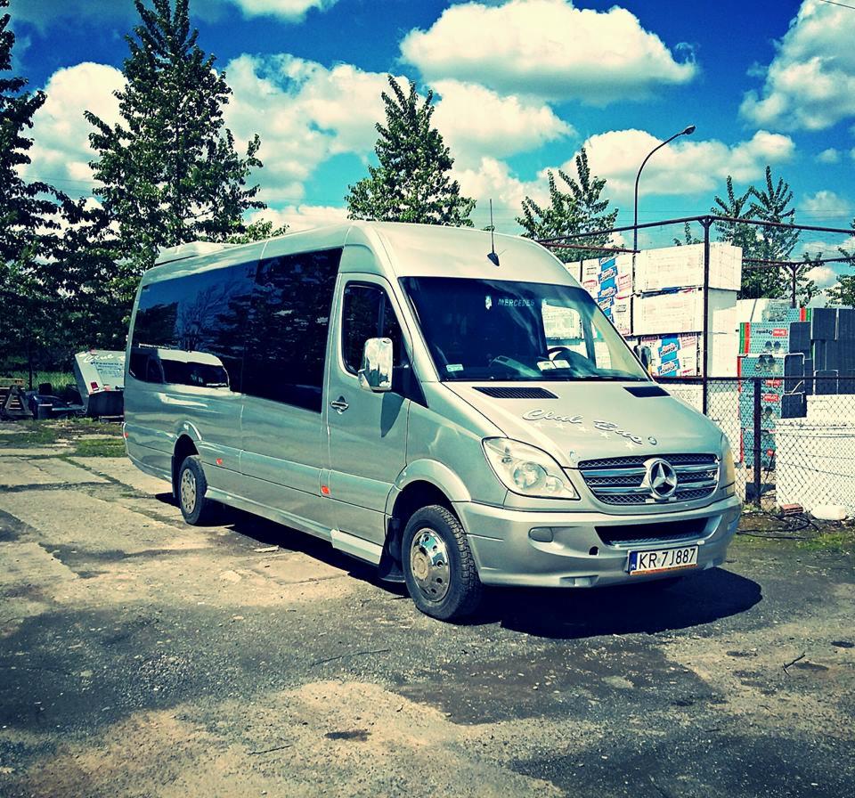 Srebrny Mercedes-Benz Sprinter z przyciemnianymi szybami, zaparkowany na betonowym placu, z widocznym napisem 'Club Bus' nad grillem i tablicą rejestracyjną KR 7J887, na tle błękitnego nieba...